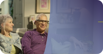 A smiling older couple sitting together on a living room couch, with a purple-blue color overlay on the right side of the image